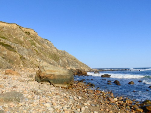 Rocky shoreline with pebbles, cliffs in the background, and gentle waves lapping at the coast under a clear blue sky.