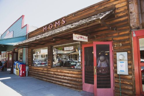 A rustic wooden storefront labeled "Moms," featuring large windows and a red door, with nearby shops visible.