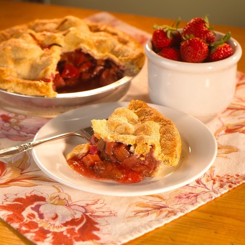 A slice of strawberry rhubarb pie on a plate, with a whole pie and fresh strawberries in a bowl nearby.