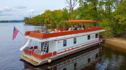 A houseboat docked by a river, surrounded by trees, with people enjoying the deck under a sunny sky.