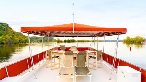 A boat deck with chairs and a table, overlooking a calm lake surrounded by green hills and trees.