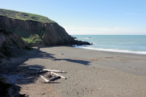 A serene beach scene with gentle waves, sandy shore, and cliffs in the background under a clear blue sky.