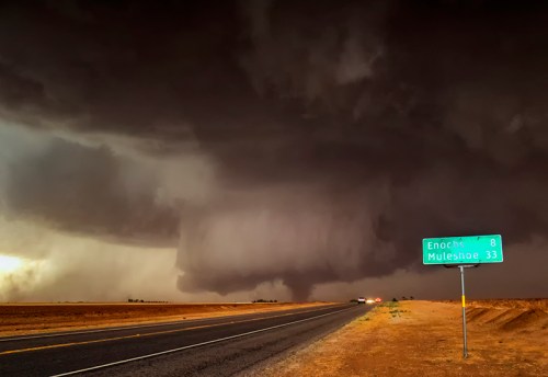 A dark, stormy sky looms over a highway with a sign indicating distances to Enochs and Muleshoe.