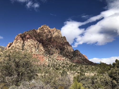 A rugged mountain rises against a blue sky with scattered clouds, surrounded by desert vegetation.