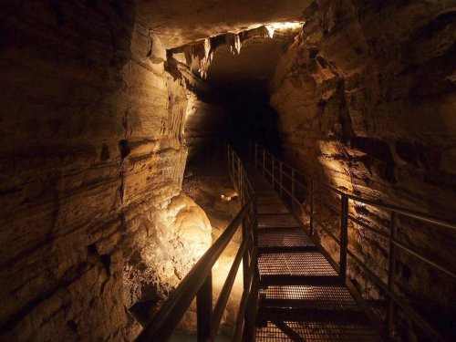 A dimly lit cave with a metal walkway, showcasing rock formations and illuminated areas along the walls.