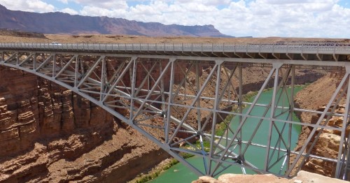 A steel bridge spans a deep canyon with a green river below and mountains in the background under a partly cloudy sky.