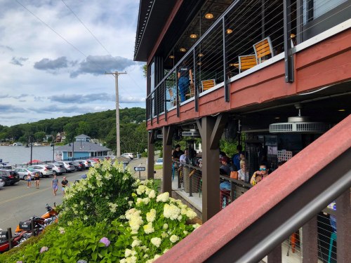 A lakeside restaurant with outdoor seating, surrounded by greenery and parked cars along the waterfront.
