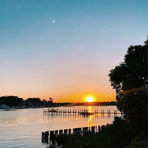 A serene sunset over a calm river, with a dock and an American flag silhouetted against the vibrant sky.