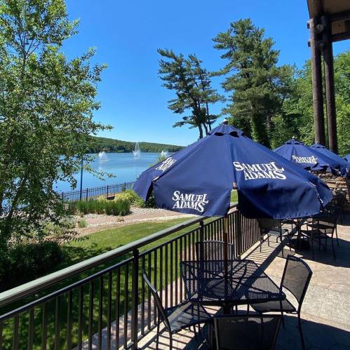 A scenic view of a lake with fountains, featuring outdoor seating under blue umbrellas with the Samuel Adams logo.