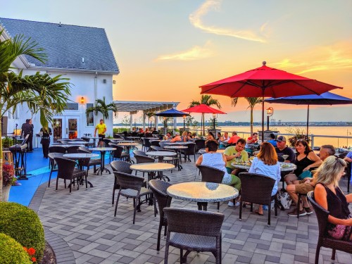 Outdoor dining area with people enjoying meals, colorful umbrellas, and a sunset view over the water.