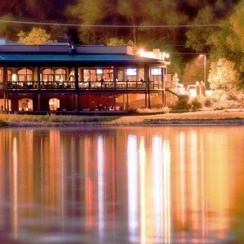 A lakeside restaurant illuminated at night, reflecting colorful lights on the water's surface.