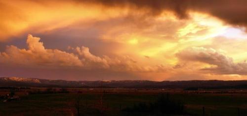 A dramatic sunset over a landscape, with vibrant orange and yellow hues illuminating the clouds and distant mountains.