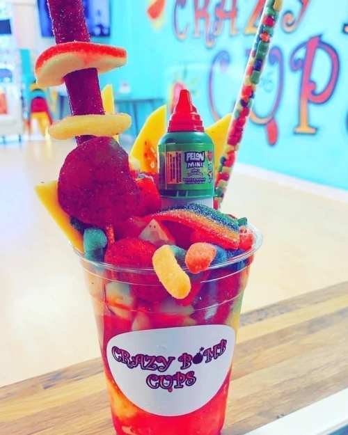 A colorful dessert cup filled with fruit, candy, and a bottle of flavored syrup, set against a vibrant background.