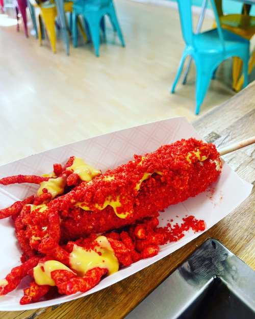 A corn dog coated in red hot Cheetos, drizzled with yellow mustard, served on a paper tray with extra Cheetos.