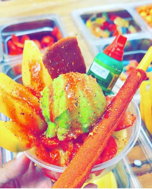A colorful fruit cup with mango, watermelon, and green ice cream, topped with chili powder and a tamarind stick.