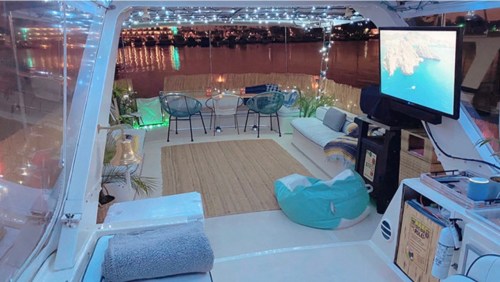 Cozy boat interior with string lights, seating area, and a TV, overlooking a calm waterfront at night.