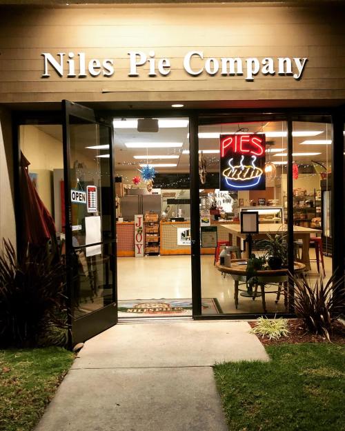 Exterior of Niles Pie Company with an "OPEN" sign and a bright "PIES" neon sign, inviting customers inside.