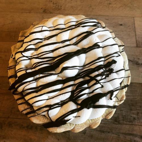A round pie topped with fluffy white meringue and drizzled with chocolate sauce on a wooden surface.