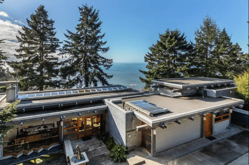 Modern house with solar panels, surrounded by trees, overlooking a scenic ocean view under a clear blue sky.