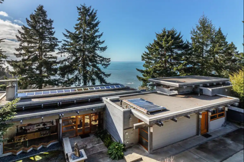 Modern house with solar panels, surrounded by trees, overlooking a scenic ocean view under a clear blue sky.