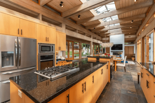 Modern kitchen with wooden cabinets, granite countertops, and large windows, featuring a central island and stainless appliances.