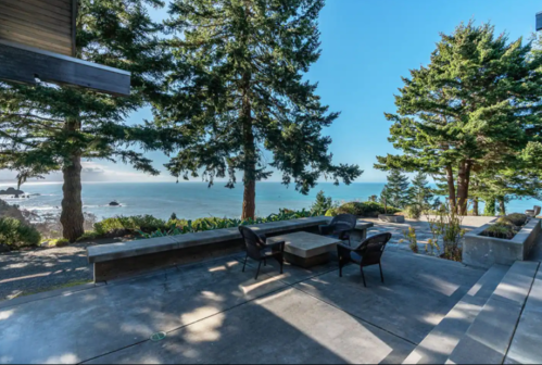 A scenic outdoor patio with chairs, overlooking a tranquil ocean view framed by tall trees.