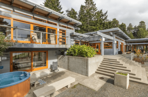 Modern building with large windows, a hot tub, and landscaped steps, surrounded by trees and a cloudy sky.