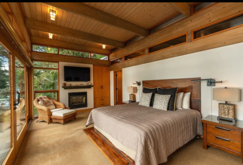 Modern bedroom with a large bed, wooden accents, a cozy chair, and large windows overlooking a natural view.