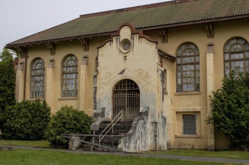 Abandoned building with a weathered facade, large windows, and a staircase leading to a gated entrance.