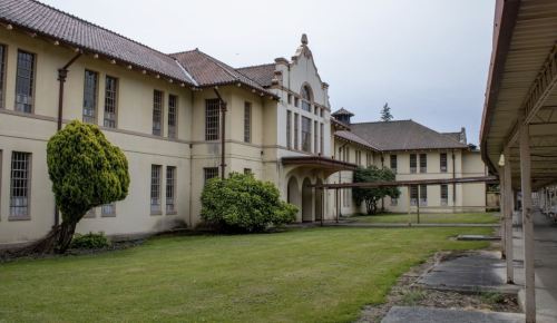 An old, two-story building with a tiled roof, surrounded by grass and shrubs, under a cloudy sky.