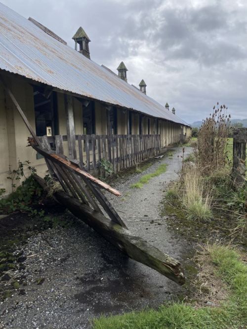 A weathered building with a damaged fence and overgrown path, under a cloudy sky.