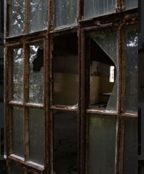 A close-up of a broken window with shattered glass and rusted metal frames, revealing a dimly lit interior.