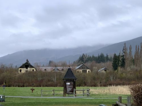 Abandoned buildings surrounded by trees and grass, with mountains and cloudy skies in the background.