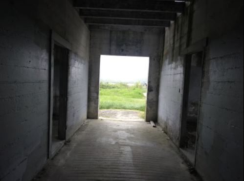 A dimly lit corridor with concrete walls, leading to an open doorway and a view of green fields outside.