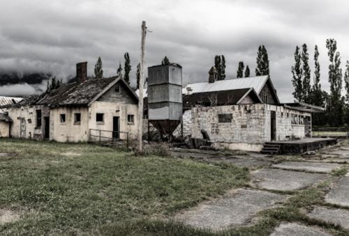 Abandoned buildings with weathered exteriors, overgrown grass, and a cloudy sky in the background.