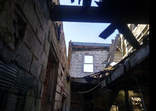 View of a dilapidated building interior, showing exposed beams and a window against a clear blue sky.