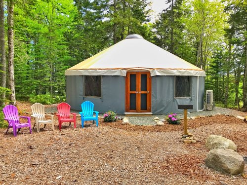 A yurt surrounded by trees, with colorful chairs in front and a small garden area.