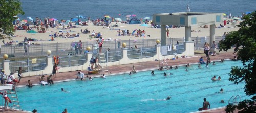 A busy outdoor swimming pool with people swimming, lifeguards on duty, and a beach with umbrellas in the background.