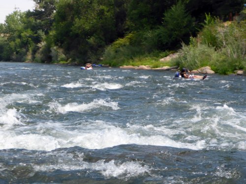 Two people float on tubes down a river, surrounded by lush greenery and rocky banks. Waves ripple in the water.