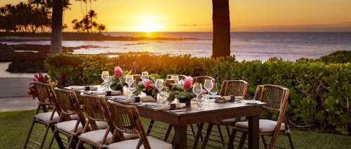 A beautifully set outdoor dining table at sunset, overlooking the ocean with lush greenery and tropical flowers.