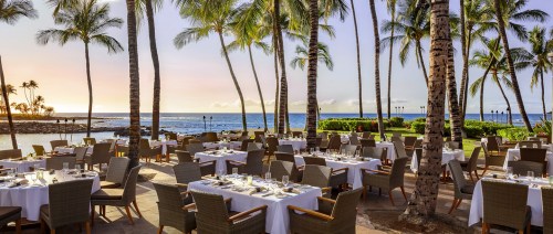 Outdoor dining area with tables set for a meal, surrounded by palm trees and a view of the ocean at sunset.