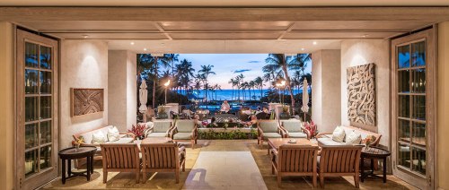 A luxurious lounge area with wooden furniture, overlooking a tropical beach at sunset, framed by palm trees.