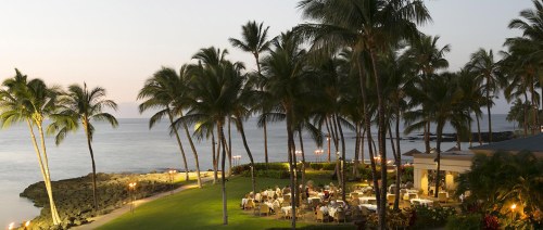A serene beachfront dining area surrounded by palm trees at dusk, with tables set for guests.