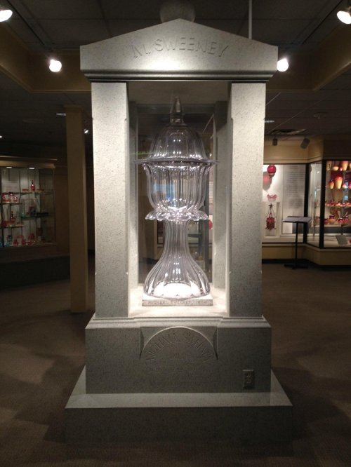 A glass sculpture displayed in a museum, encased in a stone structure with soft lighting and surrounding exhibits.