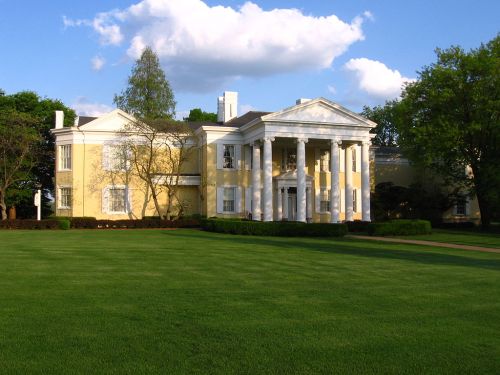 A large yellow mansion with white columns, surrounded by green grass and trees under a blue sky with fluffy clouds.