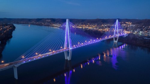 Aerial view of a illuminated cable-stayed bridge over a river at dusk, with city lights in the background.