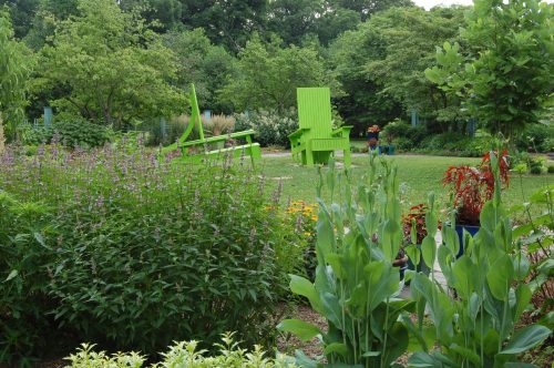 A vibrant garden featuring green chairs, lush plants, and colorful flowers under a clear sky.
