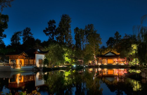 A serene night scene of a tranquil garden with illuminated buildings reflecting in a calm pond.
