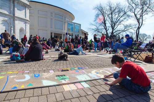 A vibrant outdoor scene with people creating chalk art on pavement, surrounded by a large crowd and trees.