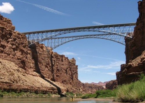 A large steel bridge spans a canyon, with rocky cliffs and a river below under a clear blue sky.